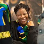 Eine Frau mit geflochtenem Haar lächelt freundlich und trägt einen blauen, grünen, gelben und schwarzen Schal. Sie sitzt in einem Innenraum, und ein Teil eines Tansania-Schals ist zu sehen. Im Hintergrund ist ein verschwommenes Bild eines Löwen zu sehen.