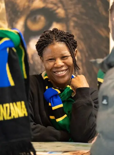 Eine Frau mit geflochtenem Haar lächelt freundlich und trägt einen blauen, grünen, gelben und schwarzen Schal. Sie sitzt in einem Innenraum, und ein Teil eines Tansania-Schals ist zu sehen. Im Hintergrund ist ein verschwommenes Bild eines Löwen zu sehen.