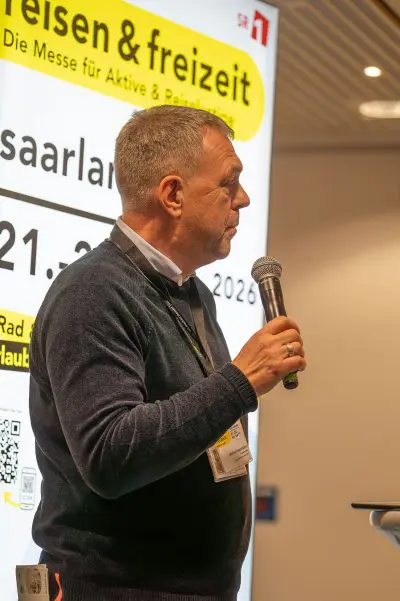 Ein Mann mit einem Mikrofon spricht auf einer Veranstaltung vor einem großen Display mit deutschem Text über eine Reise- und Freizeitmesse, einschließlich Daten und Ortsangaben.