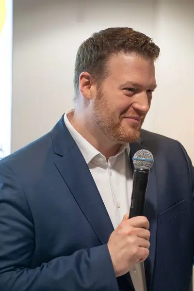 Ein Mann in blauem Anzug und weißem Hemd hält lächelnd ein Mikrofon in der Hand und scheint bei einer Veranstaltung in einem Gebäude zu sprechen.