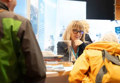 Eine lächelnde Frau mit Brille sitzt an einem Stand und unterhält sich mit zwei Personen. Auf dem Tisch liegen ein Schiffsmodell und Broschüren. Die Umgebung scheint eine Ausstellung oder Veranstaltung zu sein.