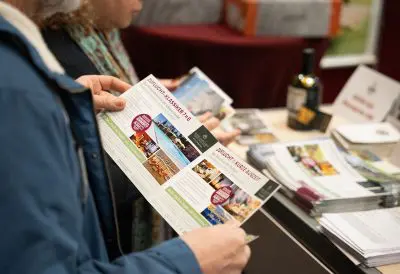 Eine Person, die an einem Stand eine Broschüre hält, mit einer weiteren Person an ihrer Seite. Auf dem Tisch liegen Broschüren, Zeitschriften und eine Flasche, was auf eine Werbe- oder Informationsveranstaltung hindeutet.