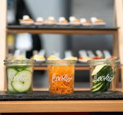 Drei Gläser mit der Aufschrift Cookit enthalten geschnittene Gurken, geraspelte Karotten und weitere Gurkenscheiben, die auf einer schwarzen Fläche mit unscharfen Vorspeisen auf einem Ständer im Hintergrund ausgestellt sind.