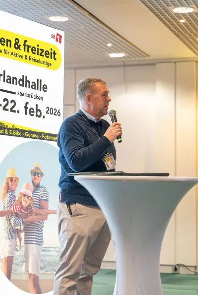 Ein Mann steht an einem runden Tisch und spricht bei einer Indoor-Veranstaltung in ein Mikrofon. Hinter ihm ist ein leuchtend gelbes Werbebanner mit einem Text über eine Freizeitmesse in Saarbrücken angebracht, auf dem eine Familie am Strand abgebildet ist.