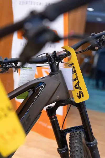Nahaufnahme eines schwarzen Fahrrads mit einem gelben Schild mit der Aufschrift SALE! am Lenker, das in einem Innenraum mit unscharfen Schildern und Menschen im Hintergrund ausgestellt ist.