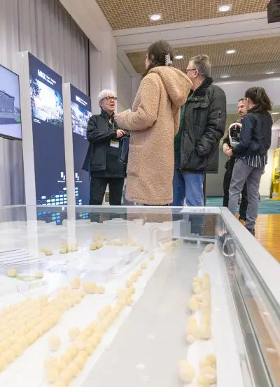 Mehrere Personen stehen und unterhalten sich in einem Ausstellungsraum, mit großen Schautafeln im Hintergrund. Im Vordergrund befindet sich ein glasüberdachtes Architekturmodell mit kleinen gelben Bäumen.