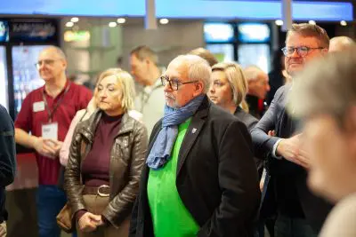 Eine Gruppe Erwachsener, sowohl Männer als auch Frauen, steht in einem Haus und hört aufmerksam zu. Im Mittelpunkt steht ein älterer Mann mit einer Brille, einem grünen Pullover und einem blauen Schal. Im Hintergrund sind helle Lichter und unscharfe Menschen zu sehen.