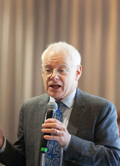 Ein älterer Mann mit Brille und weißem Haar, der einen Anzug und eine gemusterte blaue Krawatte trägt, spricht in ein Mikrofon. Der Hintergrund ist mit vertikalen Linien verschwommen.