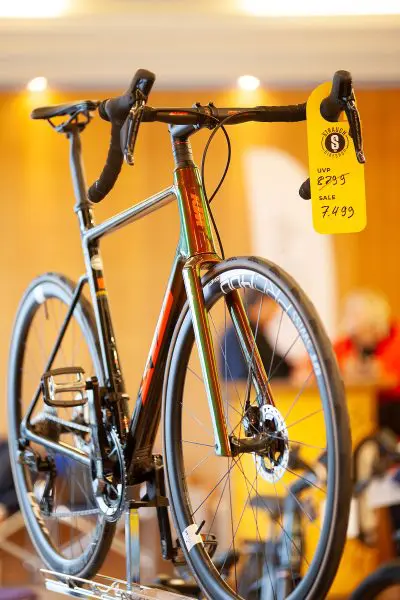 Ein Rennrad wird in einem Innenraum mit einem gelben Verkaufsschild ausgestellt, auf dem der ursprüngliche Preis von 8.295 € und ein Verkaufspreis von 7.499 € angegeben sind. Der Hintergrund ist leicht unscharf und hebt das Fahrrad hervor.