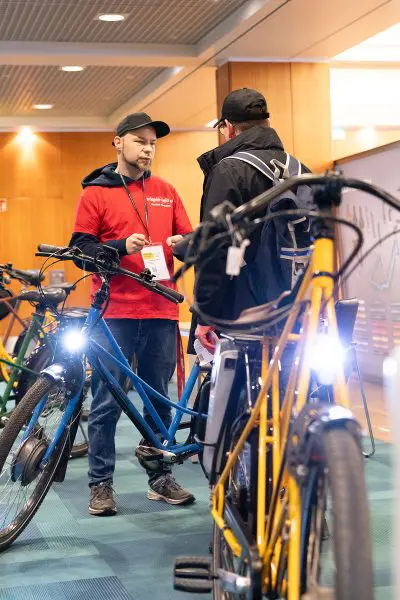Ein Mann in einem roten Hemd unterhält sich mit einem anderen Mann in einer schwarzen Jacke neben Fahrrädern in einem Innenraum. Beide stehen in einem gut beleuchteten Raum mit Holzwänden neben einem blauen und einem gelben Fahrrad, die hell beleuchtet sind.
