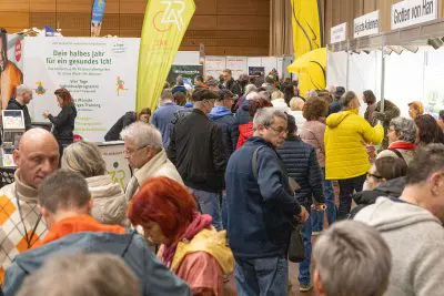 Eine überfüllte Indoor-Messe mit Menschen, die herumlaufen und Stände besuchen, einige lesen Schilder und Broschüren. Mehrere Banner und Informationsdisplays sind zu sehen, mit heller Beleuchtung und einer geschäftigen Atmosphäre.