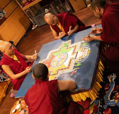 Vier buddhistische Mönche in roten Roben arbeiten gemeinsam an einem farbenfrohen Sandmandala, wobei sie sorgfältig Sand auf eine große ebene Fläche in einem Raum mit Holzböden und Regalen im Hintergrund legen.