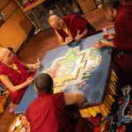 Vier Mönche in roten Roben sitzen um einen Tisch herum und gestalten mit großer Sorgfalt ein farbenfrohes Sandmandala mit komplizierten geometrischen Mustern und Symbolen. Der Schauplatz scheint ein Innenraum zu sein, in dem sich verschiedene Materialien befinden.