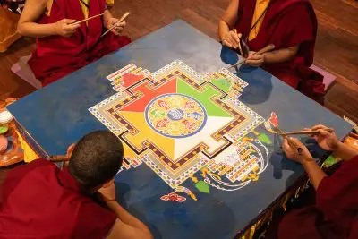 Vier Mönche in roten Roben sitzen um einen Tisch und gestalten mit dünnen Metallwerkzeugen sorgfältig ein buntes Sandmandala mit komplizierten geometrischen Mustern auf einer blauen Oberfläche.