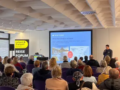 Ein Redner hält einen Vortrag vor einem Publikum in einem Reiseforum. Auf einem Bildschirm werden Informationen über eine Kreuzfahrt nach Norwegen und Island angezeigt, und es ist ein Schild "reisen & freizeit" zu sehen. Die Zuhörer sitzen mit Blick auf die Präsentation.