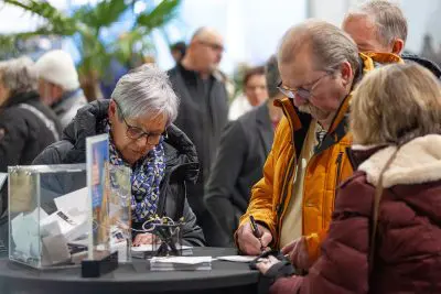 Eine Gruppe älterer Erwachsener steht um einen Tisch herum, füllt Formulare aus und schreibt. Es scheint sich um eine Veranstaltung in einem Gebäude zu handeln, bei der im Hintergrund weitere Personen zu sehen sind.
