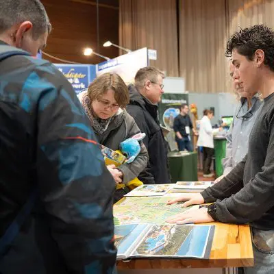 Menschen versammeln sich um einen Stand bei einer Veranstaltung in einer Halle. Eine Frau, die ein blaues Stofftier in der Hand hält, prüft Broschüren auf einem Tisch, während andere zuschauen und zwei Personen hinter dem Stand stehen und sich mit den Besuchern unterhalten.