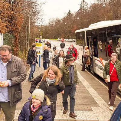 Eine Gruppe von Menschen, darunter Erwachsene und Kinder, geht an einem bewölkten Tag einen Bürgersteig entlang und steigt aus einem Bus aus. Einige Menschen sind in Jacken und Schals gehüllt. Im Hintergrund sind Bäume und eine Straße zu sehen.