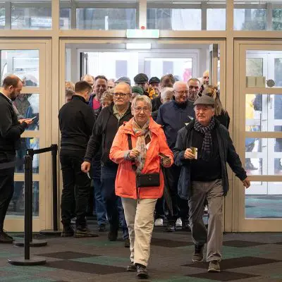 Eine Gruppe älterer Menschen, viele mit Jacken und Hüten, geht durch den Eingang eines Gebäudes. Zwei Personen führen die Gruppe an, lächeln und halten Getränke in der Hand, andere folgen ihnen.
