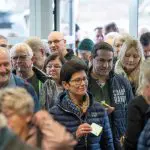 Eine Gruppe von Menschen, sowohl Männer als auch Frauen, steht in einem Innenraum dicht beieinander in einer Warteschlange. Die meisten tragen Jacken, einige halten Papiere oder Eintrittskarten in der Hand und scheinen auf ein Ereignis oder einen Eingang zu warten.