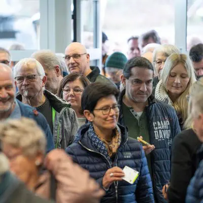Eine Gruppe von Menschen, sowohl Männer als auch Frauen, steht in einem Innenraum dicht beieinander in einer Warteschlange. Die meisten tragen Jacken, einige halten Papiere oder Eintrittskarten in der Hand und scheinen auf ein Ereignis oder einen Eingang zu warten.