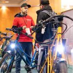 Zwei Männer stehen in einer Halle zwischen mehreren Fahrrädern. Der eine, der ein rotes Hemd und eine Mütze trägt, spricht mit dem anderen, der schwarz gekleidet ist. Die Scheinwerfer der Fahrräder leuchten hell im Vordergrund.