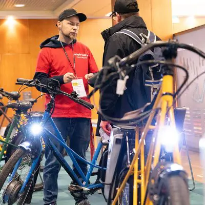Zwei Männer stehen in einer Halle zwischen mehreren Fahrrädern. Der eine, der ein rotes Hemd und eine Mütze trägt, spricht mit dem anderen, der schwarz gekleidet ist. Die Scheinwerfer der Fahrräder leuchten hell im Vordergrund.