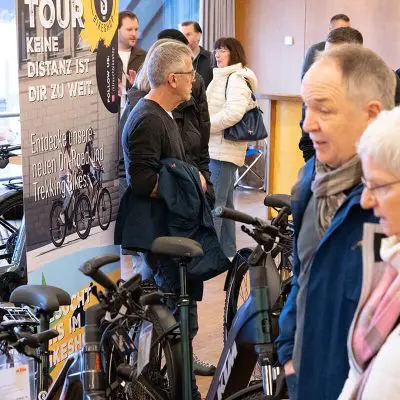 Eine Gruppe von Menschen steht in einem Innenraum auf einer Fahrradmesse und betrachtet Fahrräder und Auslagen. Im Hintergrund sind Banner mit deutschen Texten und Fahrradbildern zu sehen.