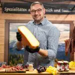 Ein lächelnder Mann mit Brille und schwarzen Handschuhen hält einen großen Käseblock an einem Marktstand mit Wurst, Weintrauben, Gläsern und Fleischproben vor dem Hintergrund von Bergen und einem Holzschild in deutscher Sprache.