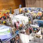 Eine belebte überdachte Ausstellungshalle mit zahlreichen Ständen und Auslagen, die von Menschen bevölkert sind, die herumlaufen, sich unterhalten und die verschiedenen Exponate erkunden. Die Atmosphäre wirkt lebendig und geschäftig.