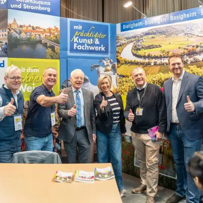 Sechs lächelnde Menschen stehen in einem Innenraum an einem Veranstaltungsstand zusammen und zeigen die Daumen nach oben. Hinter ihnen sind bunte Banner mit deutschen Texten und Landschaftsbildern zu sehen. Im Vordergrund steht ein Tisch mit Broschüren.