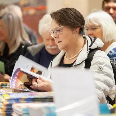 Eine Frau mit Brille und weißer Jacke liest in einer überfüllten Halle eine Zeitschrift, umgeben von anderen Erwachsenen und Stapeln von bunten Zeitschriften oder Büchern auf einem Tisch.