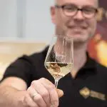 Ein lächelnder Mann mit Brille und schwarzem Hemd hält ein Glas Weißwein in die Kamera, auf dem Heimvut steht. Der Fokus liegt auf dem Weinglas, und der Mann ist im Hintergrund leicht verschwommen.