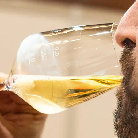 Nahaufnahme einer bärtigen Person, die ein Glas mit hellem, schaumigem Bier trinkt, das zum Mund hin gekippt ist. An der Seite des Glases sind Messmarkierungen sichtbar.