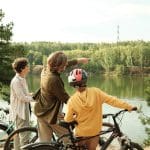 Drei Personen mit Fahrrädern stehen an einem Flussufer. Ein Erwachsener deutet auf etwas auf der anderen Seite des Wassers, während ein Kind mit Helm und ein weiterer Erwachsener in die gleiche Richtung schauen. Der ruhige Fluss ist von Bäumen umgeben.