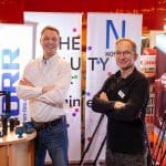 Zwei Männer stehen lächelnd mit verschränkten Armen bei einer Indoor-Veranstaltung vor bunten Bannern und Kameradisplays. Die Atmosphäre wirkt locker und freundlich.