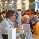 Ein lächelnder Mann hilft einem älteren Ehepaar an einem belebten Informationsschalter während einer Veranstaltung. Das Paar sieht sich Dokumente an, während im Hintergrund Menschen und Lichter verschwimmen und eine lebhafte Atmosphäre schaffen.