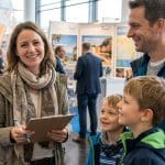 Eine Frau, die ein Klemmbrett in der Hand hält, lächelt in die Kamera, während sie sich mit einem Mann und zwei kleinen Jungen bei einer Indoor-Veranstaltung mit Informationsständen im Hintergrund unterhält.