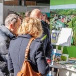 Drei Personen stehen eng beieinander in einem Innenraum und betrachten Broschüren und Hefte auf einem Schautisch. Das Sonnenlicht scheint auf sie, und im Hintergrund ist ein großes Luftbild eines grünen Campus zu sehen.