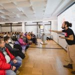 Ein Mann mit Tattoos und Bart hält in einem hellen Konferenzraum einen Vortrag vor einem sitzenden Publikum. Er gestikuliert energisch in Richtung einer projizierten Leinwand, und die Zuhörer hören aufmerksam zu.