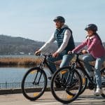 Ein Mann und ein junges Mädchen mit Helmen fahren an einem sonnigen Tag mit dem Fahrrad einen Uferweg entlang, mit Hügeln und Bäumen im Hintergrund.