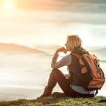 Eine Frau mit Rucksack sitzt bei Sonnenaufgang auf einem Grashügel und blickt nachdenklich auf die nebligen Berge und das sanfte Sonnenlicht in der Ferne.