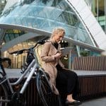 Eine Frau in einem hellbraunen Mantel sitzt im Freien neben einem Fahrrad, lächelt und benutzt ein Tablet. Sie steht vor einem modernen Glasgebäude mit kantiger Architektur.