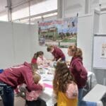 Mehrere Kinder und Erwachsene sind um einen Tisch versammelt und beschäftigen sich mit Kunst und Handwerk. Die Erwachsenen tragen passende kastanienbraune Kapuzenpullover. An der Wand hinter ihnen hängt ein buntes Banner mit Bildern von Pferden.