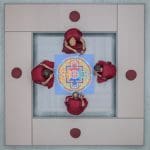 Vier Mönche in roten Roben sitzen um ein kompliziertes, farbenfrohes Mandala, das von oben betrachtet wird. Sie befinden sich in einem quadratischen, mit Kissen ausgekleideten Bereich mit vier runden roten Kissen an den Ecken.