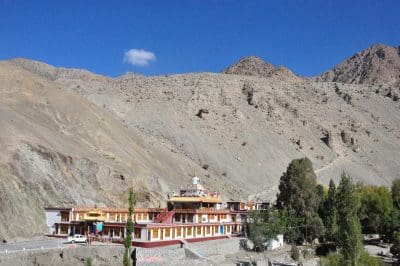Ein farbenfrohes buddhistisches Kloster mit kunstvollen Verzierungen steht am Fuße eines kargen, felsigen Berges unter einem klaren blauen Himmel, umgeben von grünen Bäumen und ein paar geparkten Fahrzeugen.