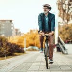 Ein lächelnder Mann mit Bart, der eine Brille, einen Helm, einen Blazer und eine braune Hose trägt, fährt mit dem Fahrrad auf einem gepflasterten Weg, der von herbstlichen Bäumen und städtischen Gebäuden gesäumt ist.