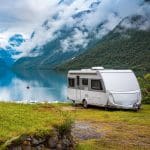 Ein weißer Wohnwagen steht auf einer Wiese am Ufer eines ruhigen Sees, umgeben von grünen Bergen mit schneebedeckten Gipfeln und dunstigen Wolken. Die friedliche Szene spiegelt sich im Wasser unter einem wolkenverhangenen Himmel.