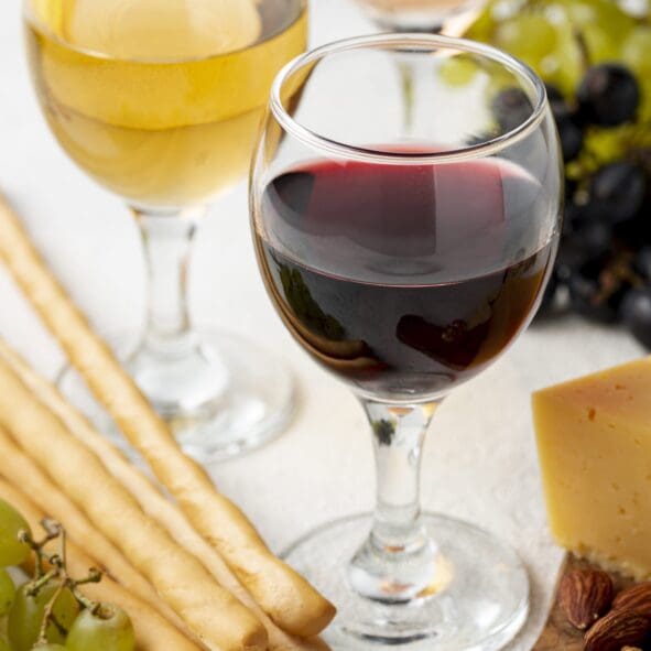 Zwei Weingläser, eines mit Rotwein und eines mit Weißwein, umgeben von Brotstangen, grünen Weintrauben, einer Käsescheibe und einigen Nüssen auf einer weißen Fläche.