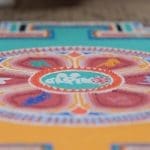 Nahaufnahme eines farbenfrohen Sandmandalas mit komplizierten kreisförmigen Mustern, einschließlich roter, grüner, oranger und gelber Formen, das auf einer flachen Oberfläche erstellt wurde. Der Fokus liegt auf der Mitte, die Ränder sind im Hintergrund verschwommen.